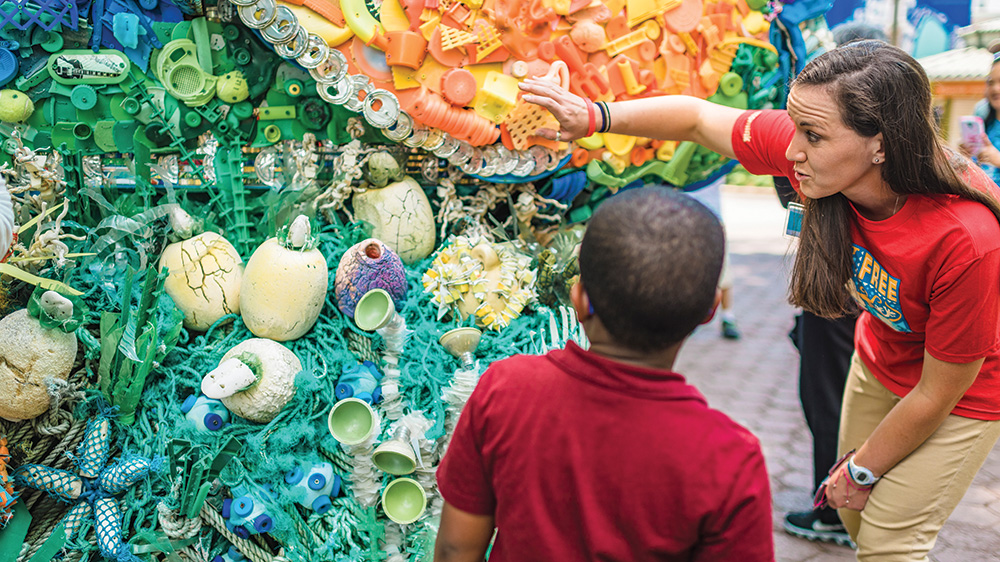 A person with pale skin and brown hair wears a red shirt and tan pants. They gesture to a colorful outdoor art display made of recycled plastic. A child with brown skin wears a red shirt and faces the display. 