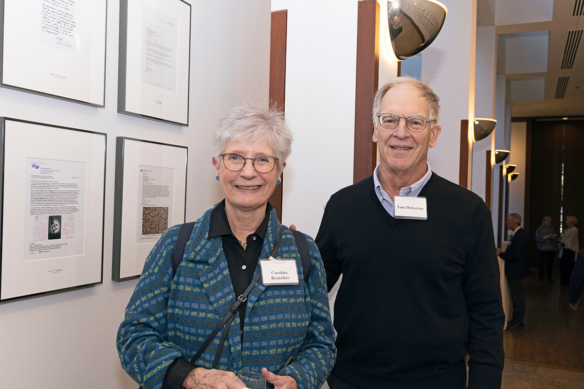 Two adults pose in front of  wall of framed letters. 