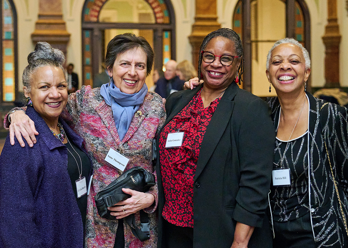 Four individuals stand shoulder to shoulder and look toward the viewer. They are smiling.
