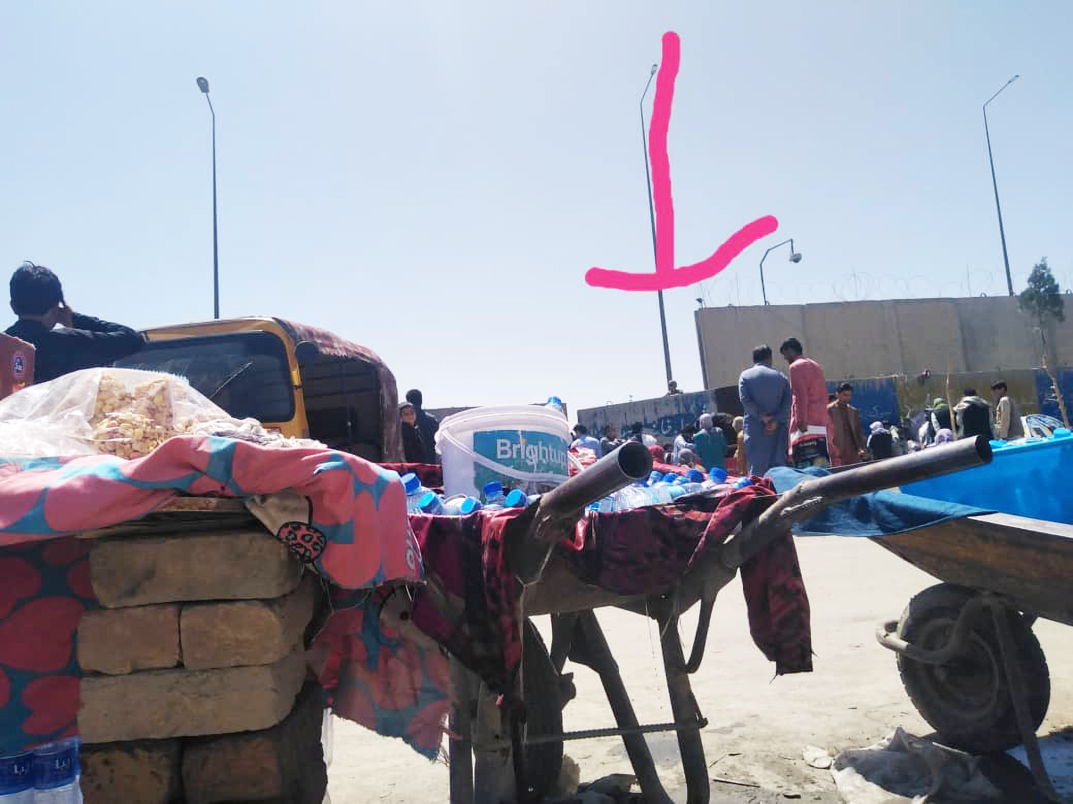 A photograph of the North Gate in Kabul, Afghanistan. Wheelbarrows and cement blocks are in the foreground, and a barrier is in the background, with a crowd behind the barrier. An arrow above the barrier points down to the center-right of the crowd.
