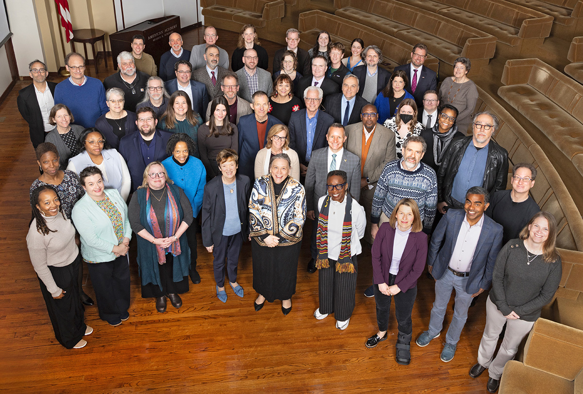 A group of about 50 adults in business casual attire pose together for a photo taken from an elevated angle with everyone smiling up at the camera. The subjects in the photo are standing in an auditorium with tan couches, wooden flooring, and white walls.