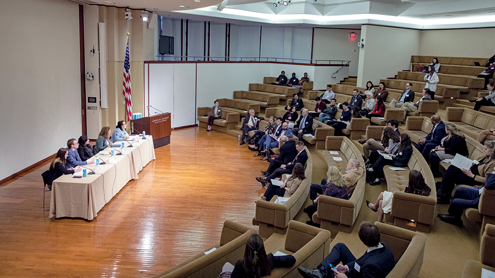 A panel of speakers sits at a table at the head of an auditorium at the American Academy of Arts and Sciences. Attendees sit in the stadium seats. 