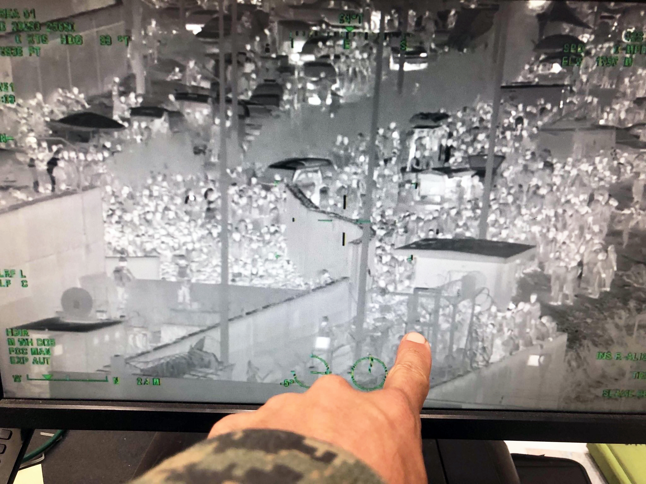 A pale hand points to a computer monitor showing an image in negative of crowds of people standing along and across the top of walls that divide sections of a town in Afghanistan. The person pointing at the monitor wears camouflage.