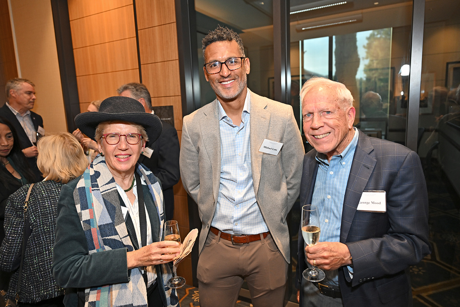 Three adults pose and smile at a crowded reception. 