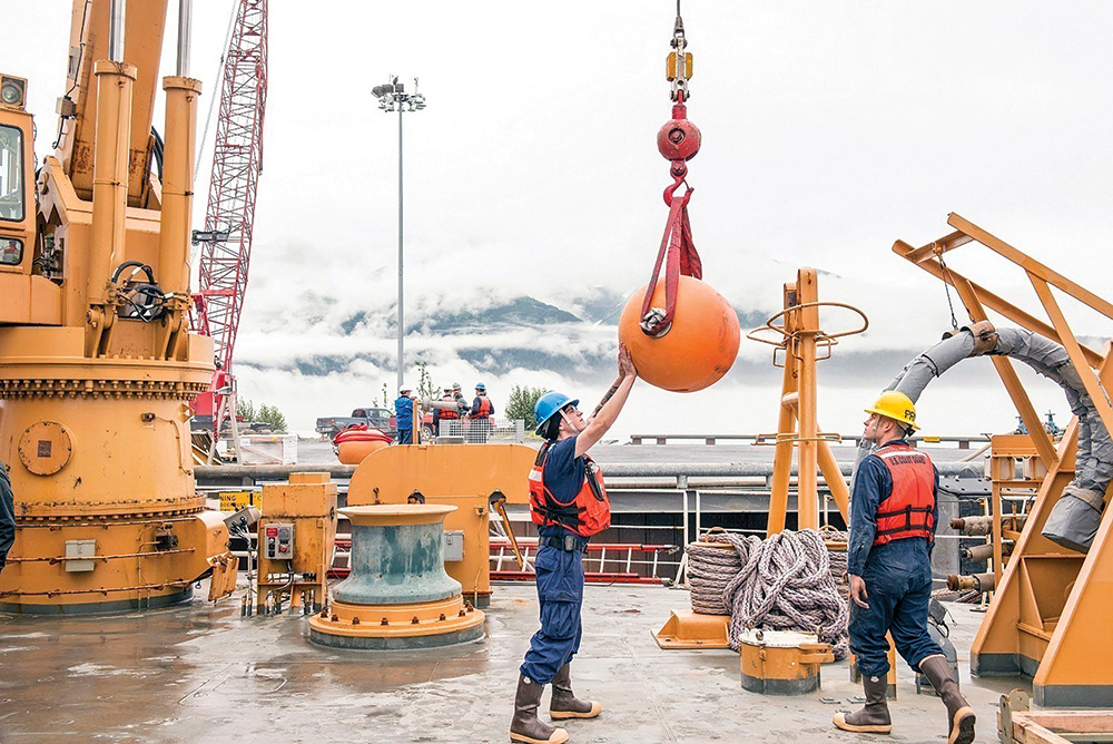 A person with pale skin wears a construction vest and hard hat and touches the bottom of an orange buoy hoisted above a ship. Another person wearing similar protective gear watches nearby.