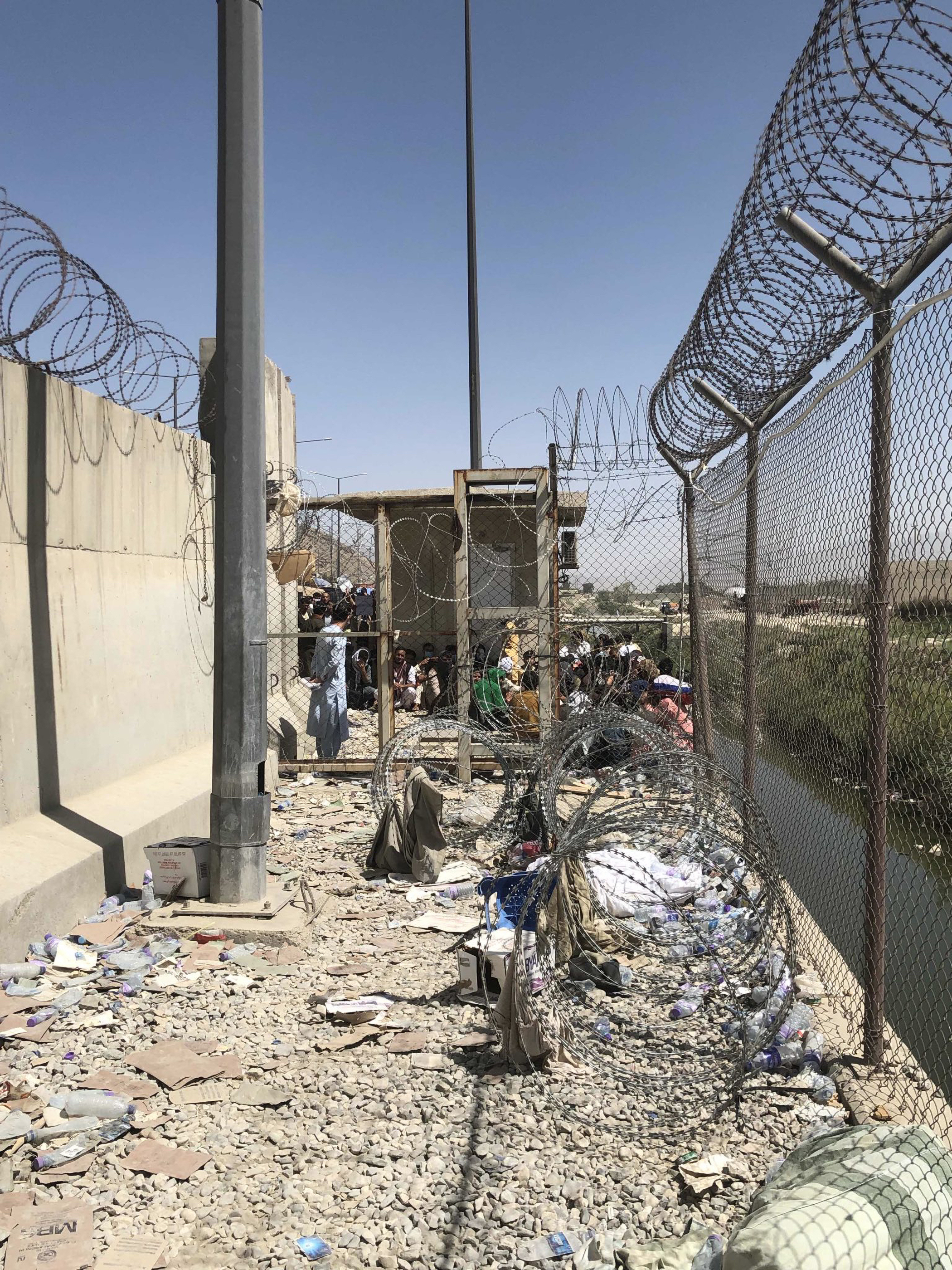 A cement expanse with a gravel floor is fenced and surrounded by barbed wire in Afghanistan. Brown people crowd the other side of the fence and face away from the camera.