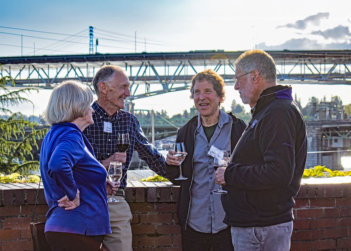 Four adults enjoy conversation in an outdoor setting with a bridge in the background.