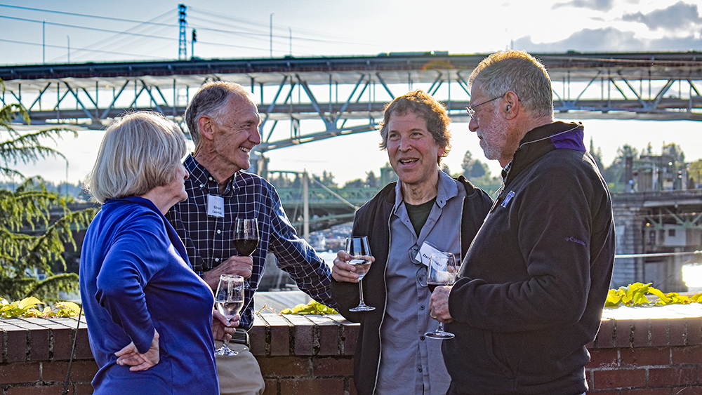 Four adults enjoy conversation in an outdoor setting with a bridge in the background.