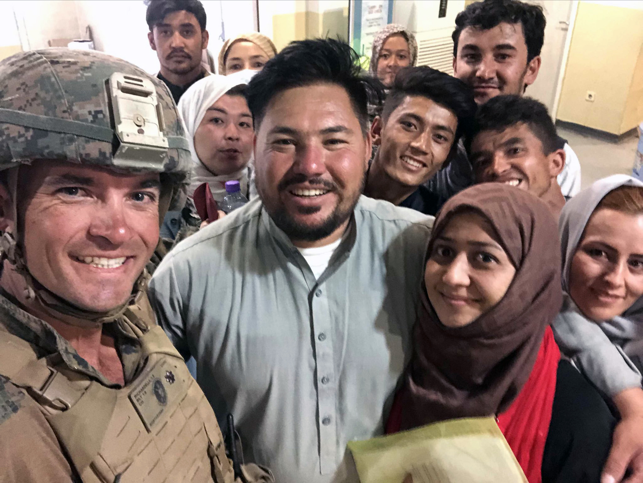 Photograph of a group of evacuees from Afghanistan standing beside a soldier. Shah and Forozan stand beside Richardella (the solider) in the foreground, and eight unnamed evacuees stand in the background. All look to the camera.