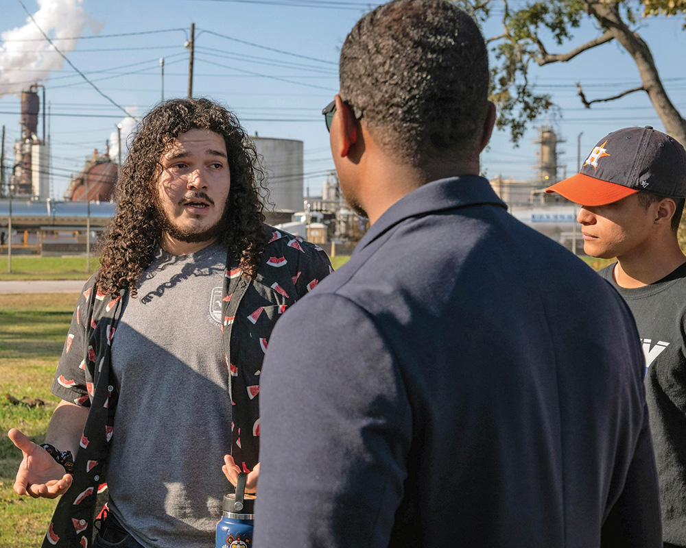 Three people stand in front of a power plant in conversation. A person with pale skin and long black curly hair faces the viewer wears casual clothes and speaks to a person with brown skin who wears a dark suit and faces away from the viewer, and a person with brown skin and short dark hair who wears casual clothes.  