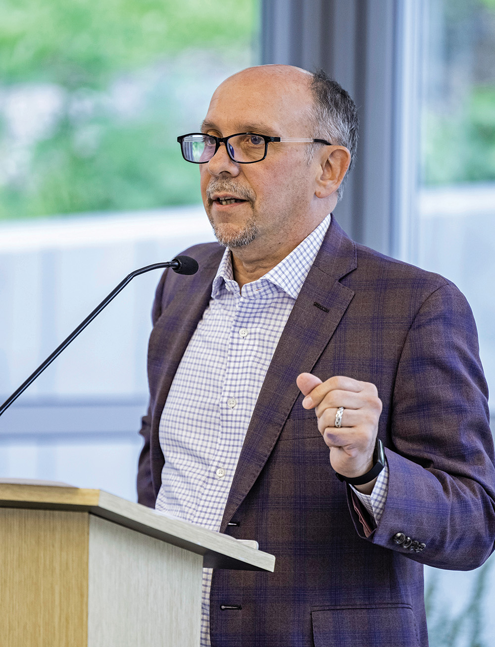 A photo of Jamie Merisotis, a person with pale skin and short graying hair. Merisotis wears a gray suit and glasses, and stands at a lectern with a microphone.