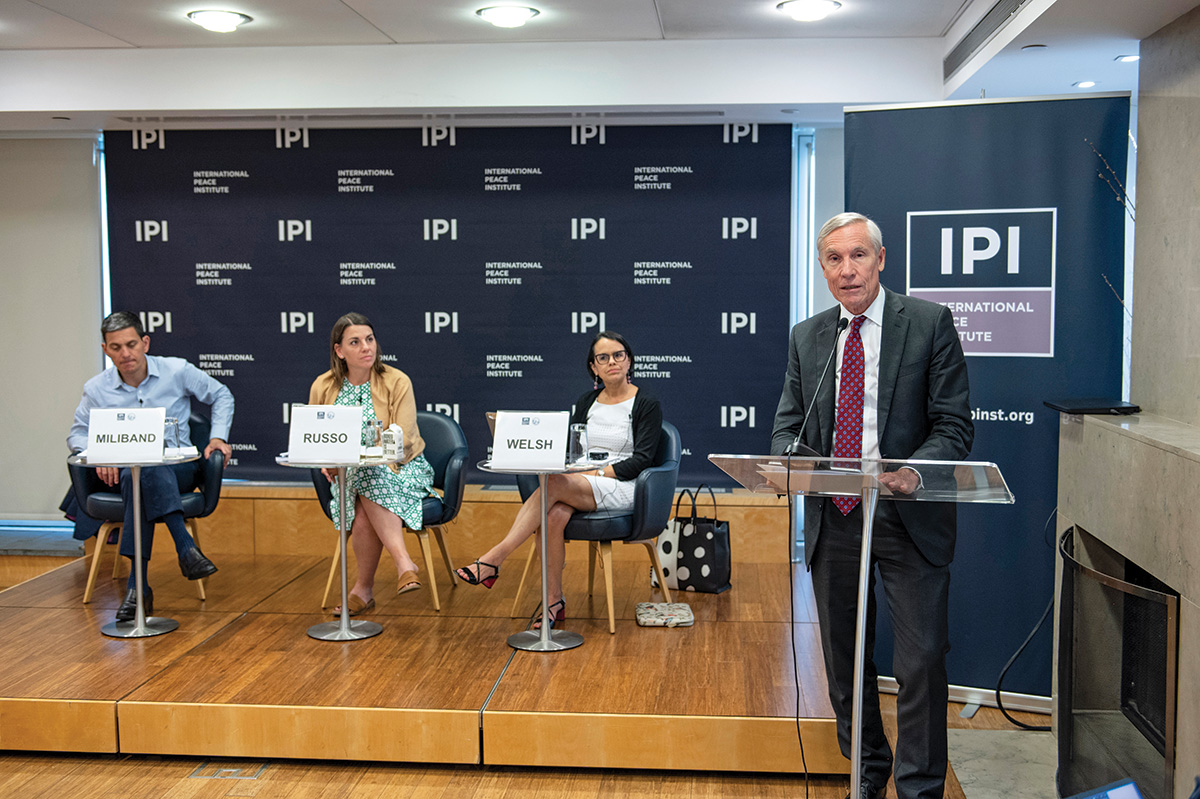 David Miliband, Jenna Russo, and Jennifer Welsh sit in front of a wall that promotes the International Peace Institute. In front of them, David Oxtoby addresses the viewer.