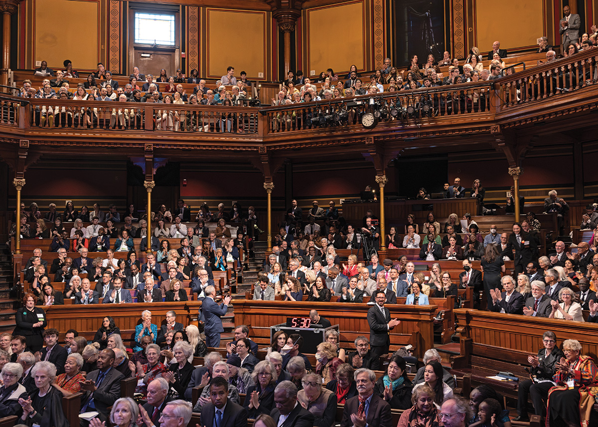 An image of the inside of Sanders Theatre at Harvard University, with people seated throughout the theatre smiling and facing the stage.