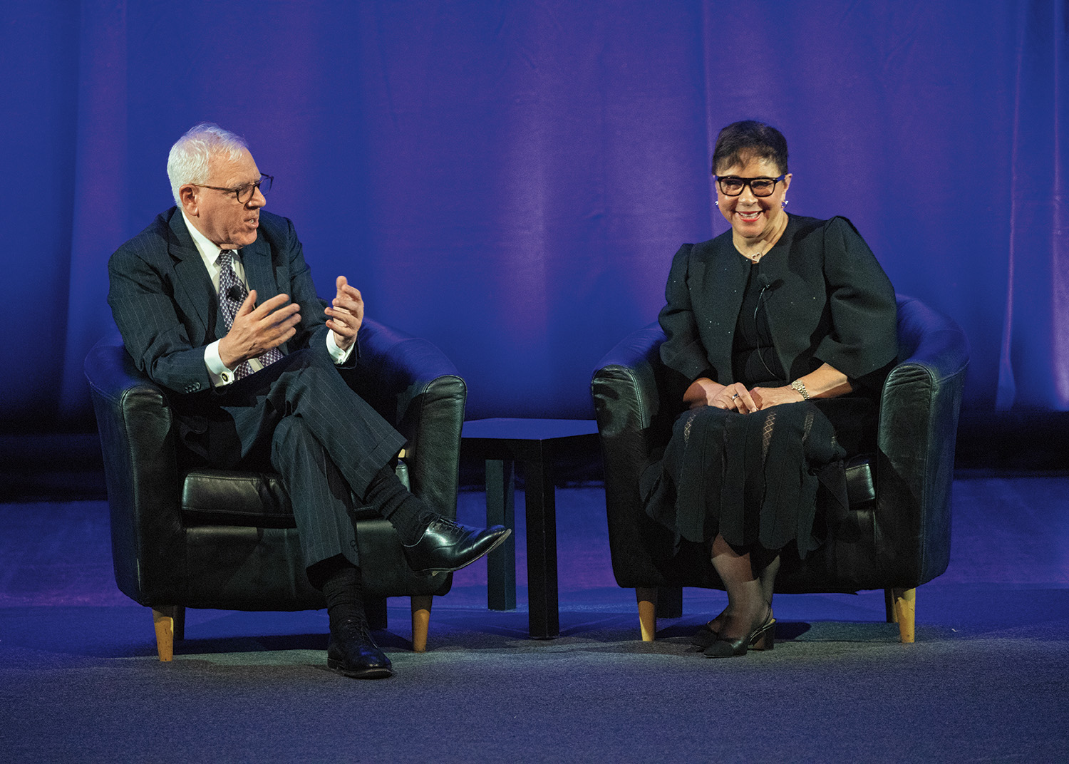 David Rubenstein and Sheila Johnson sit on a stage in large black chairs during an event at the 2023 induction ceremony. David Rubenstein wears a striped suit and glasses and has short, white hair, and pale skin. Sheila Johnson wears a black dress, coat, and glasses, and has short, black hair and light brown skin.