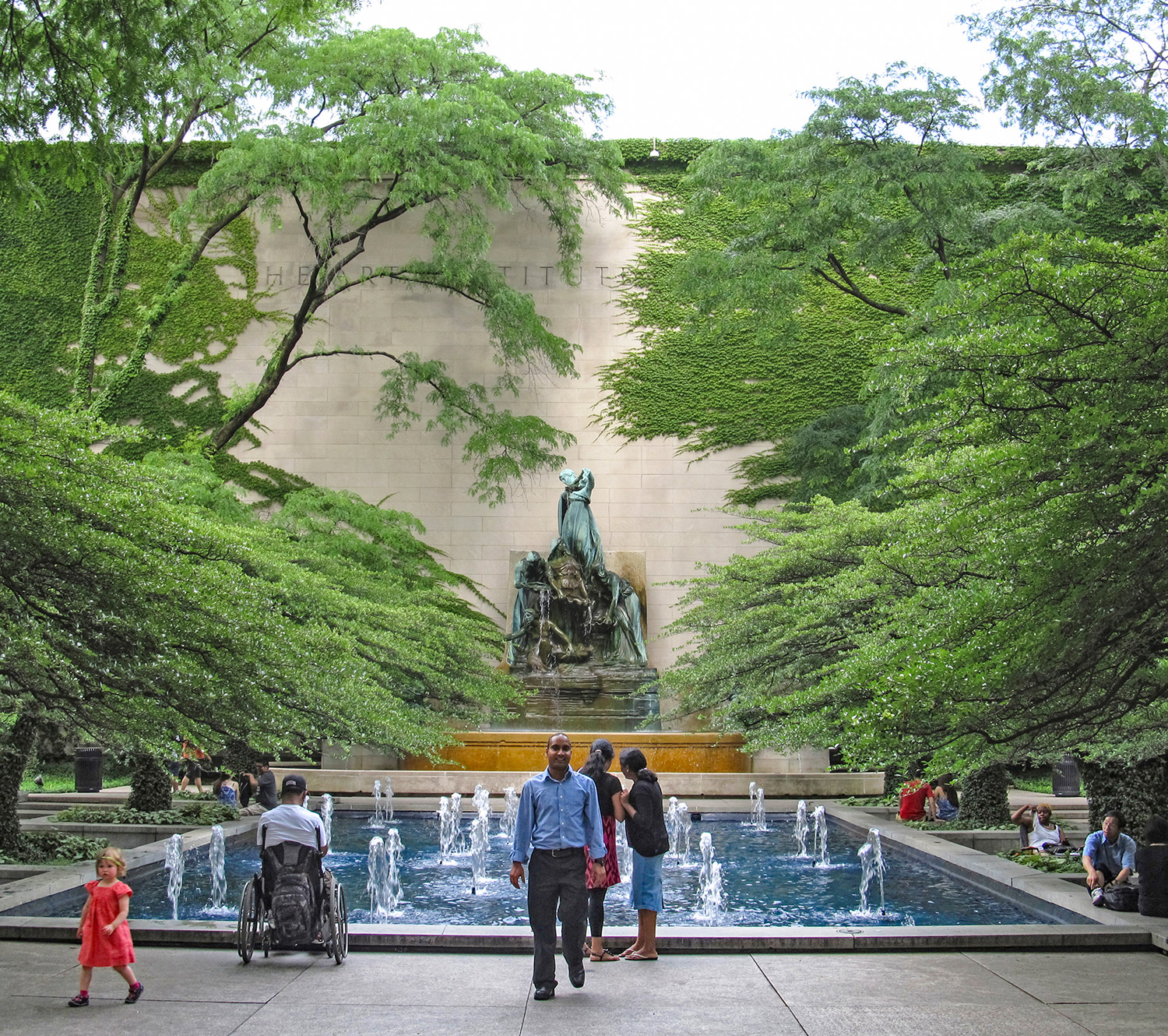 People walking and relaxing around a rectangular fountain with a statue and stone wall in the background.  The fountain is surrounded by lush green trees.