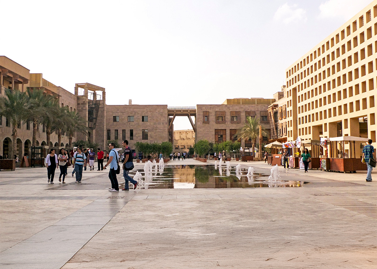 A courtyard with buildings and trees in the background and with water fountains in the center. People wearing backpacks are walking in the courtyard.