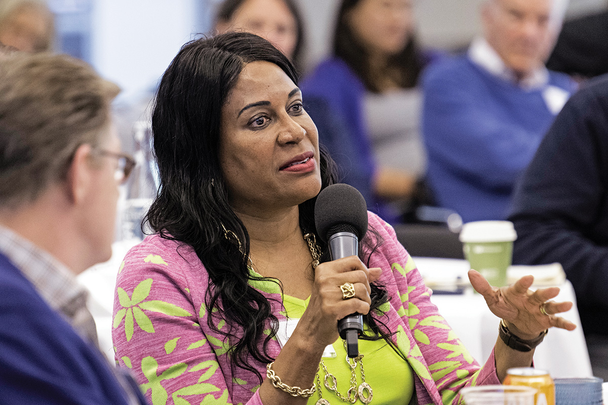 A photo of Judith Brown Clarke, a person with brown skin and long black wavy hair. Clark wears a pink and green sweater over a green blouse. She holds a microphone and speaks to a person off-camera.