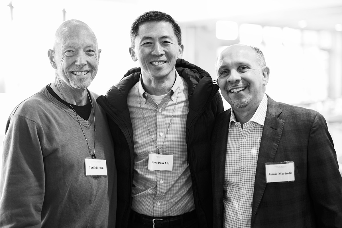 A black and white portrait of Ted Mitchell, Goodwin Liu, and Jamie Merisotis facing the camera and smiling. Mitchell has pale skin and is bald. He wears a sweater over a dark shirt. Liu has short black hair and light skin. He wears a dark coat over a checkered shirt. Merisotis has pale skin and short graying hair. He wears a gray suit and glasses.