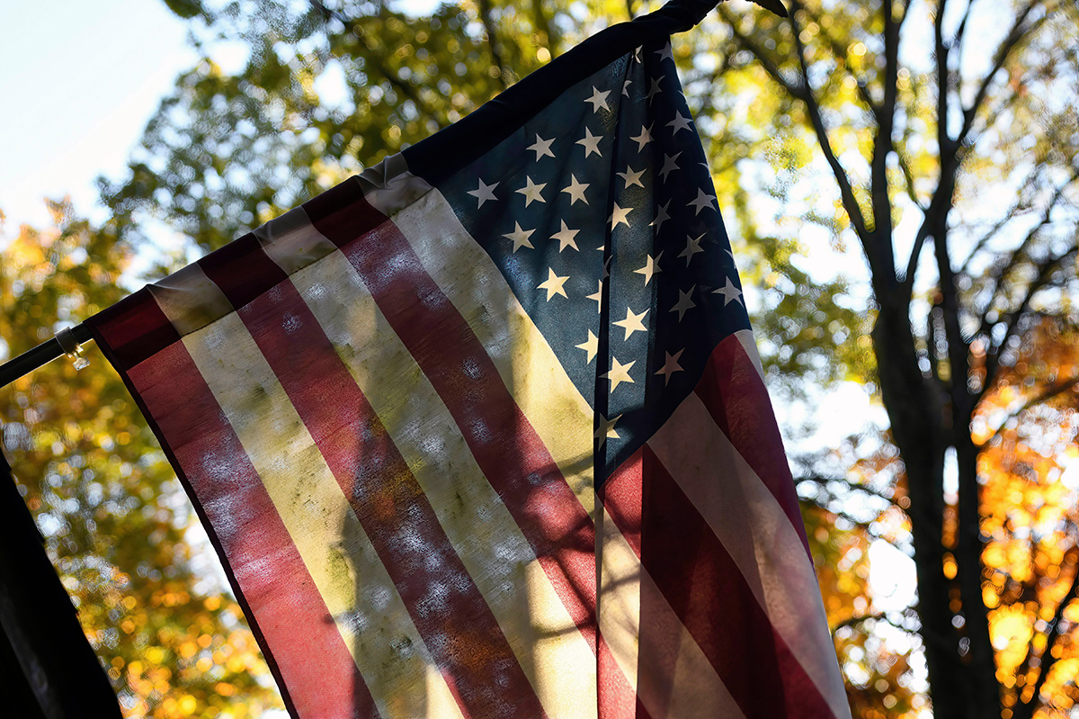 A close-up photo of the American flag with trees in the background on a sunny day.