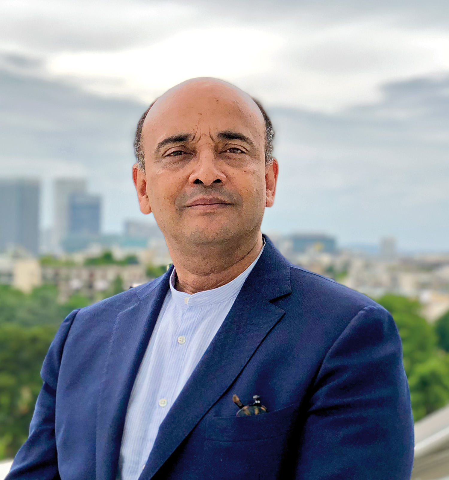 A headshot of Don M. Randel Award recipient Anthony Appiah, a person with short, dark hair and brown skin. Appiah wears a dark blue blazer and white dress shirt, with a blurry skyline view behind him. 