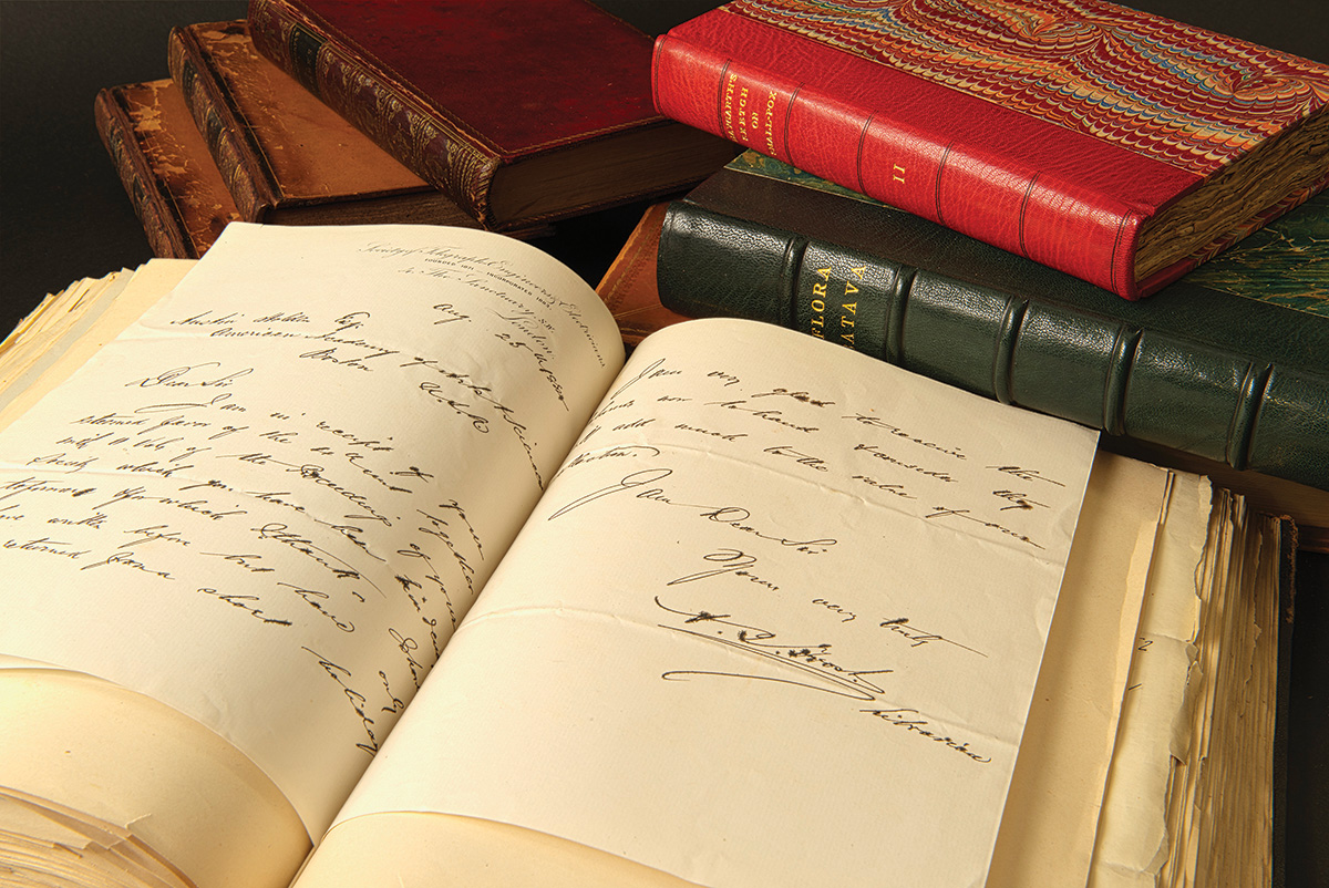 Stacks of antiquated books sit beside one open book with handwritten text on yellowing pages inside.