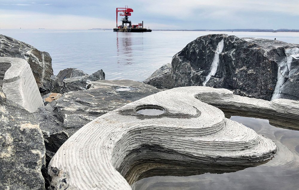 A manmade cement breakwater partially covered by water sits amid a rocky shore in the foreground. Red and gray machinery can be seen floating in the ocean in the background. 