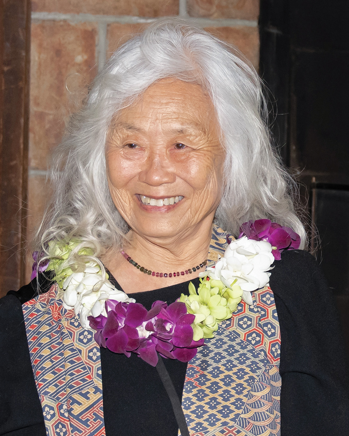 A photo of Maxine Hong Kingston, a person with brown skin and long wavy white hair. She wears a black dress under a multicolored scarf, and a necklace of white, green, and purple flowers. She looks to her right and smiles.