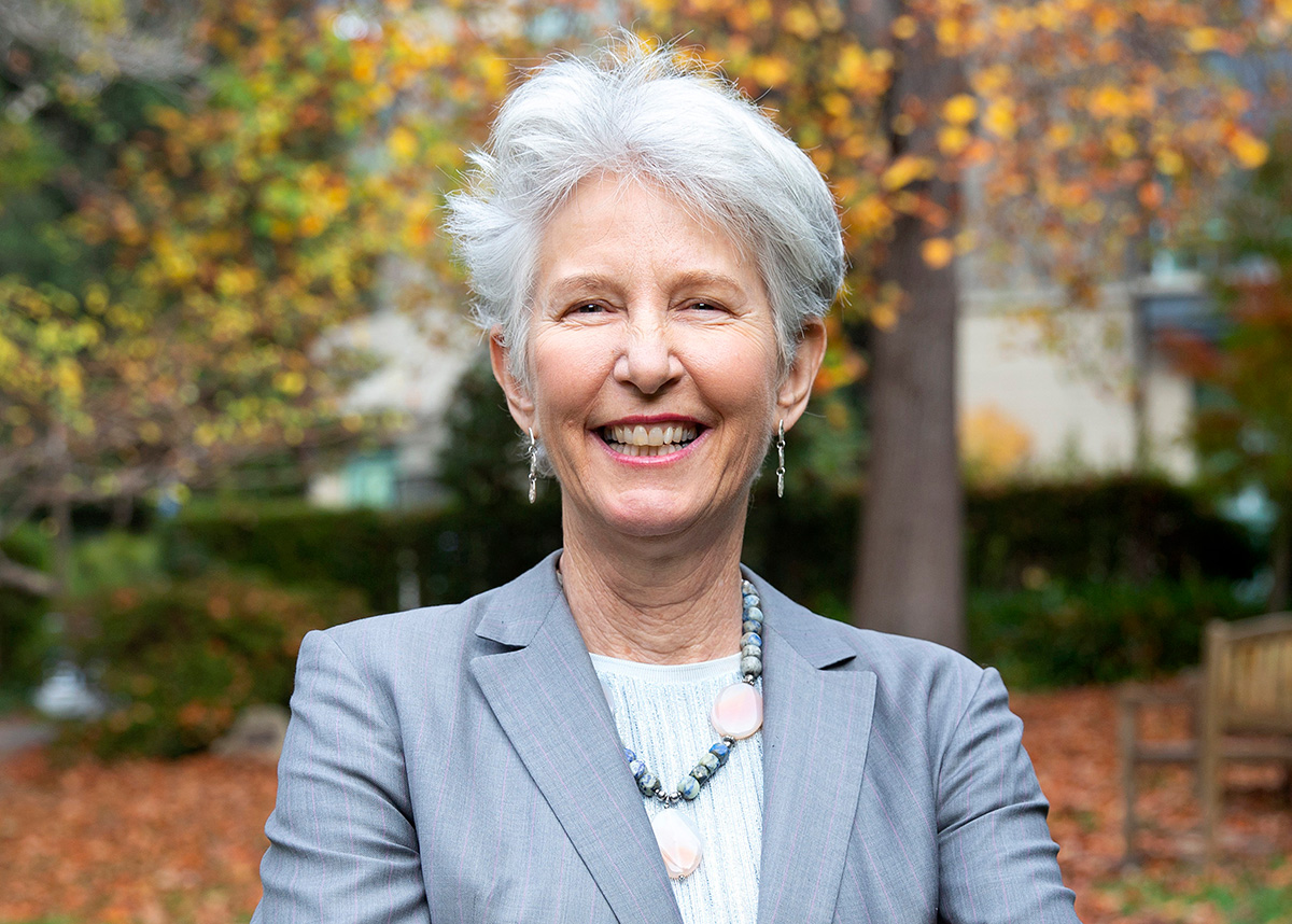 A photograph of a light-skinned individual with short gray hair.  They are wearing a light blue blazer and their arms are crossed over their chest. They are smiling at the viewer. 