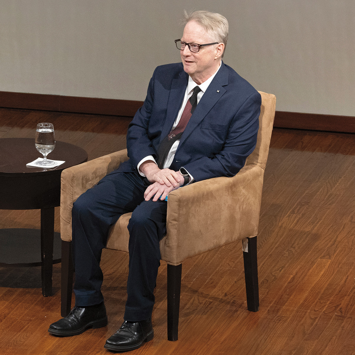 A man and a woman are sitting on chairs, engaged in a conversation. The woman is wearing a blue dress and the man is wearing a suit. There are two wine glasses on the table between them.