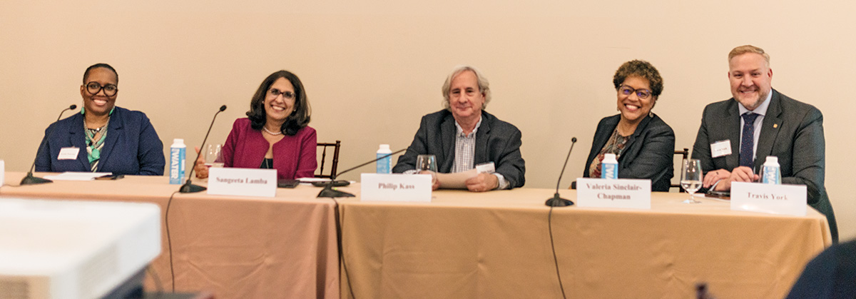 Five speakers at the event to discuss the future of higher education sit together at a long table, face the viewer and smile. 