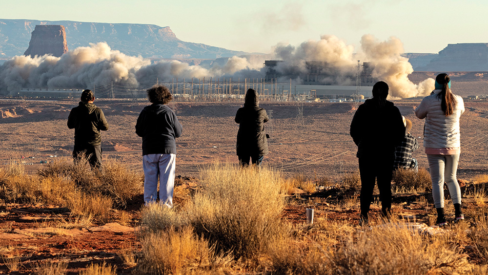 Six people stand in a desert environment with their backs to the viewer, watching a power station that is emitting smoke. Mesas are visible in the background.  