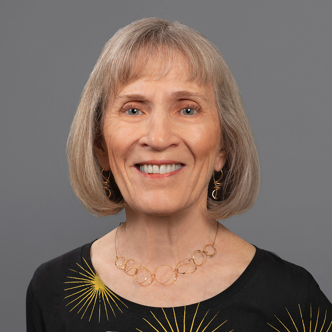 A person with short, light-colored hair is wearing a black top with a white bird and yellow starburst pattern and a gold necklace with interconnected circular links.