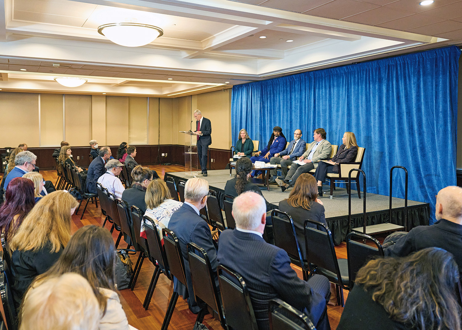 A crowd of people sitting in front of a raised stage. On the stage, one man stands at a podium, and the panelists sit.