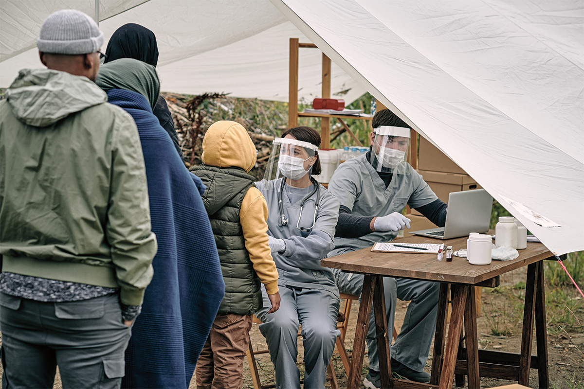 Two health care workers wearing face shields and surgical masks face a line of people waiting for medical attention. Above them, a white tarp serves as a tent.