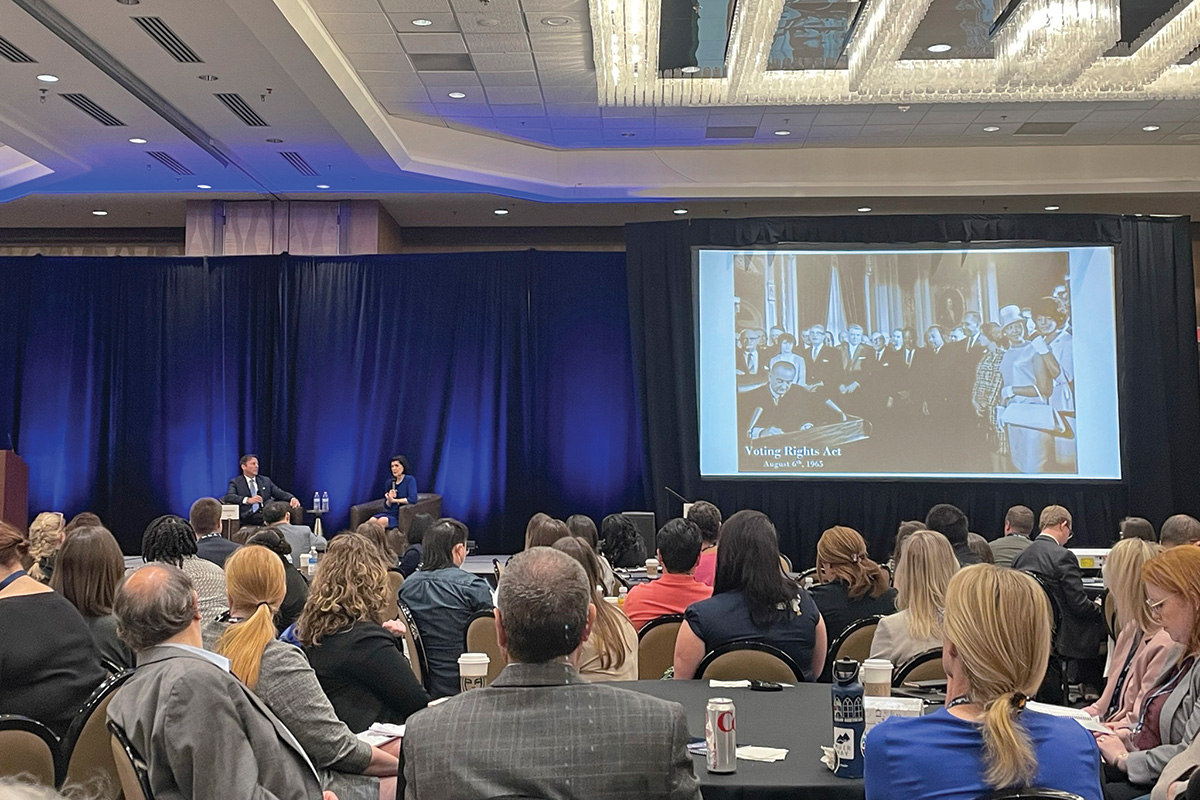 A view from a full audience of a stage with two speakers at the Equal Justice Conference. Above the stage, a screen displays a projected photo of President Lyndon Johnson signing the Voting Rights Act.