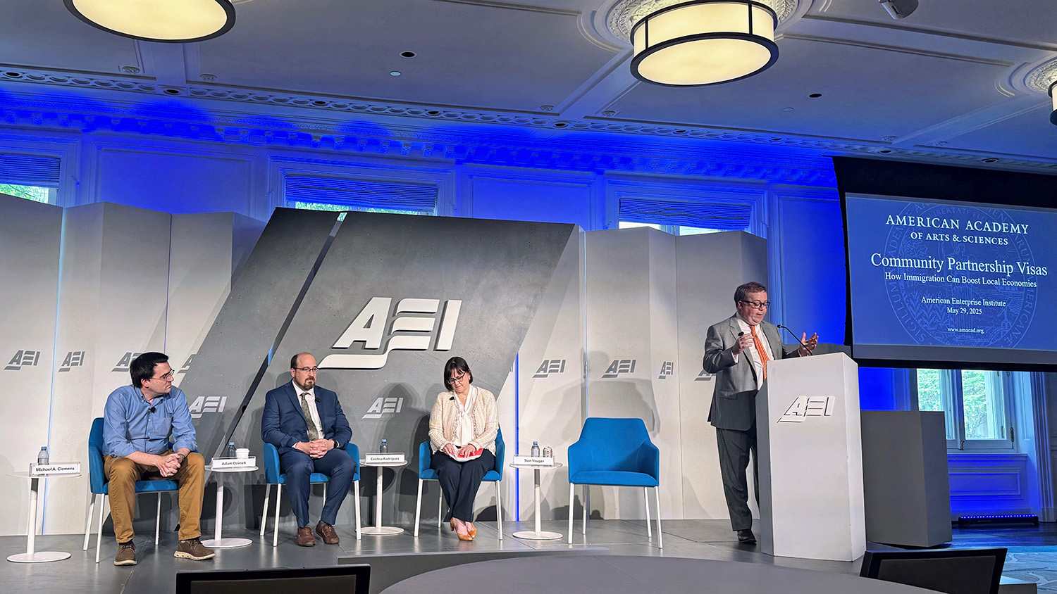 Three people sit in chairs on a stage, with a fourth person standing to their right and speaking at a microphone.  The letters AEI are on the backdrop of the stage. 