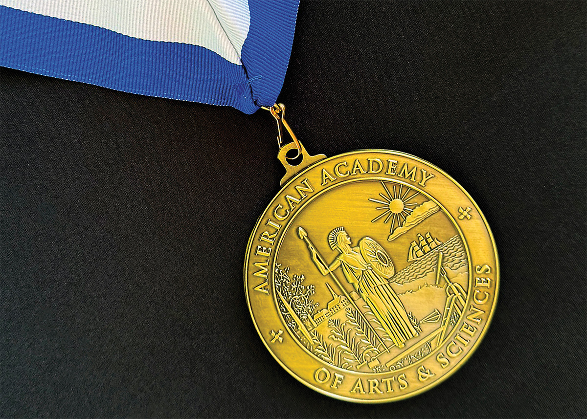A bronze circular medal, with American Academy of Arts and Sciences around the outer edges and in the center is the Roman goddess Minerva. At her right hand is a field of corn with oak trees in the background. At her left hand is the sea with a ship steering toward the town and the sun rising above a cloud. A telescope and farm tools are at her feet.