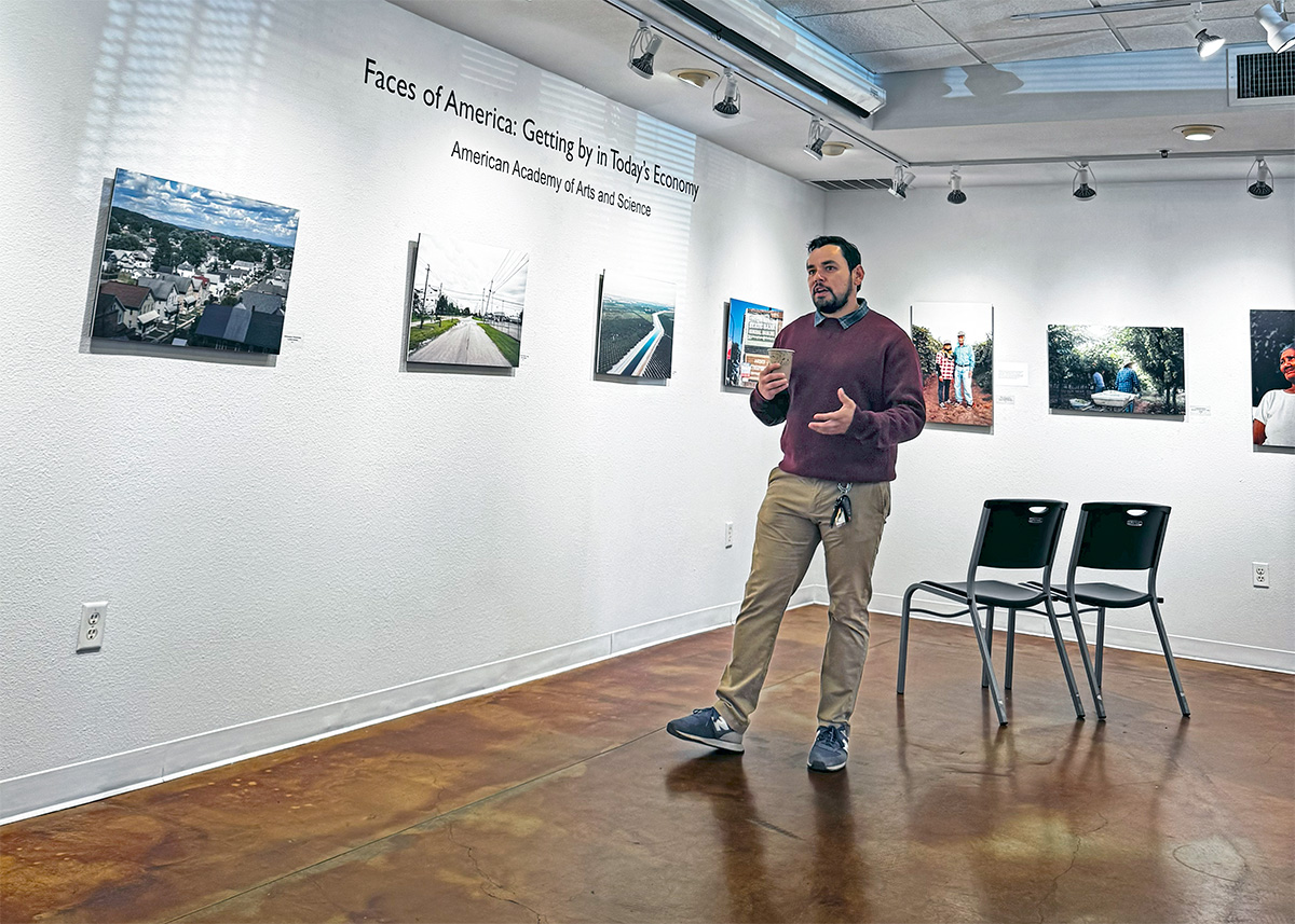 A man stands in a gallery space, in front of an exhibit entitled Faces of America: Getting by in Today’s Economy. On the walls are photographs of different communities, including aerial views of homes and an empty street as well as people in a field.  