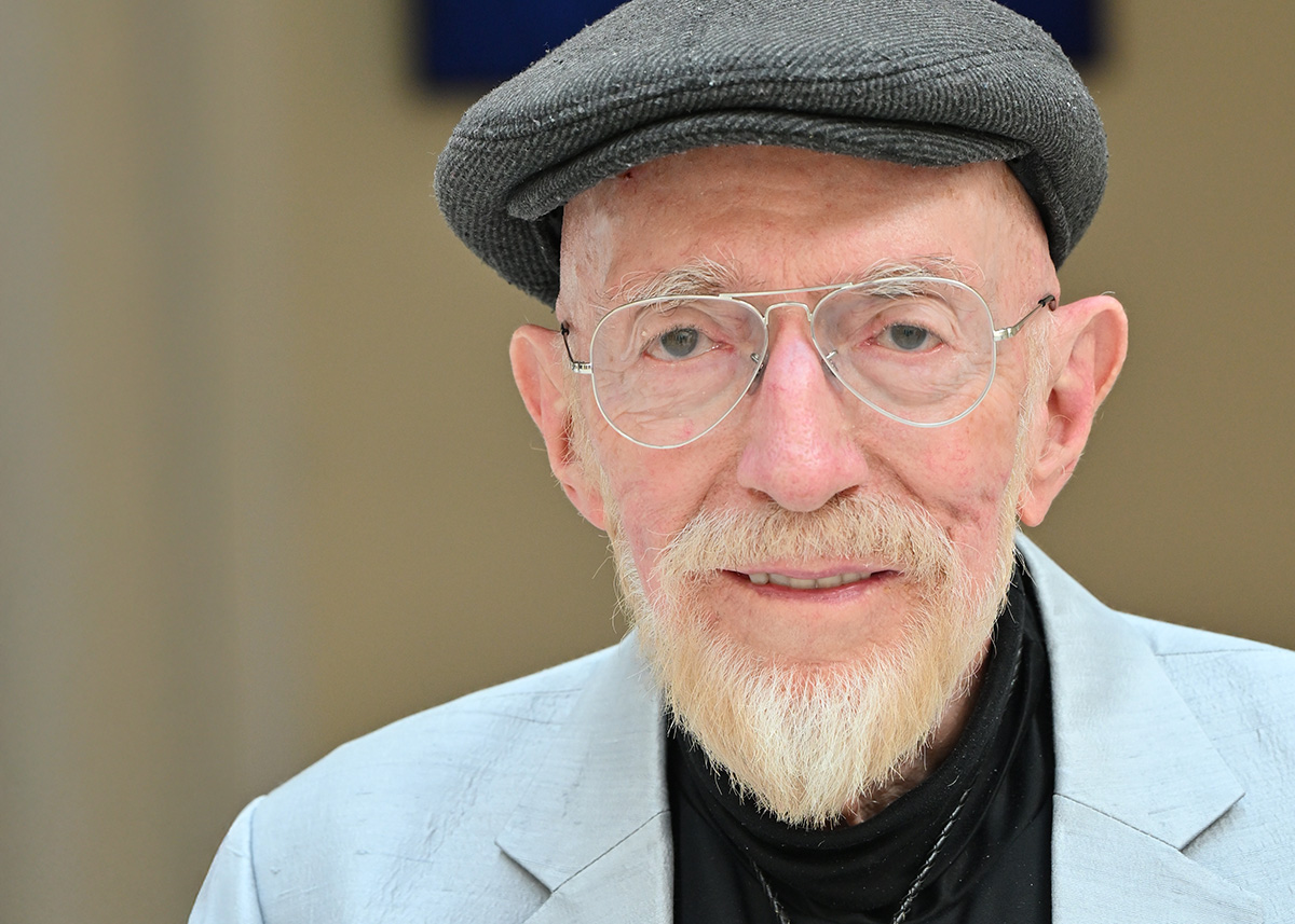 A photograph of a light-skinned man with a white beard and mustache. He is wearing a cap, glasses, a light blue sports jacket, dark shirt, and a bolo tie with a green stone. He is looking toward the viewer. 