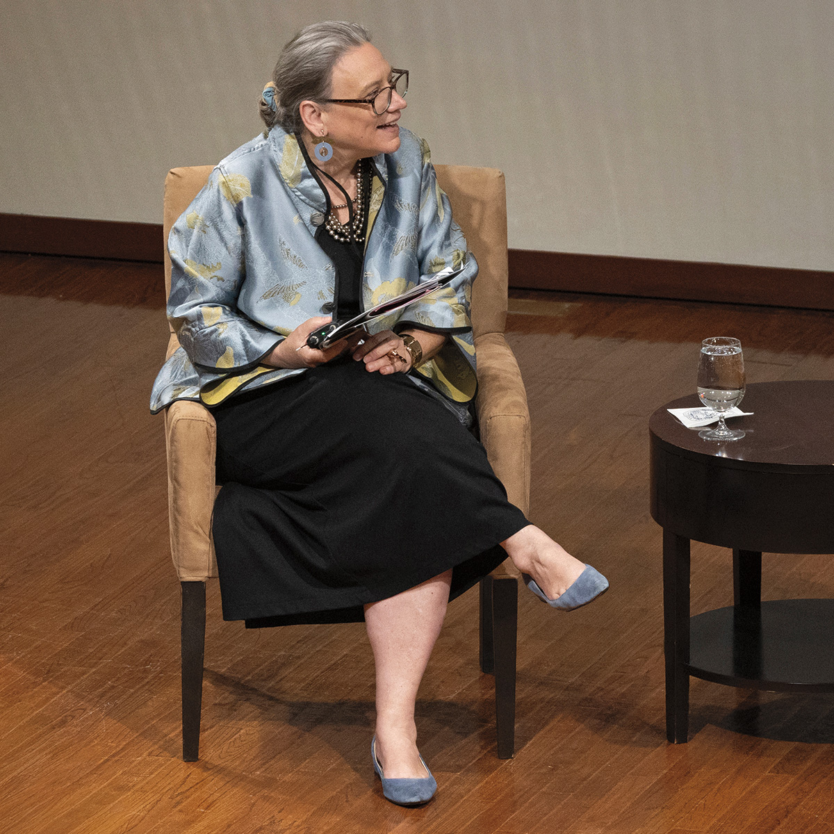 A man and a woman are sitting on chairs, engaged in a conversation. The woman is wearing a blue dress and the man is wearing a suit. There are two wine glasses on the table between them.