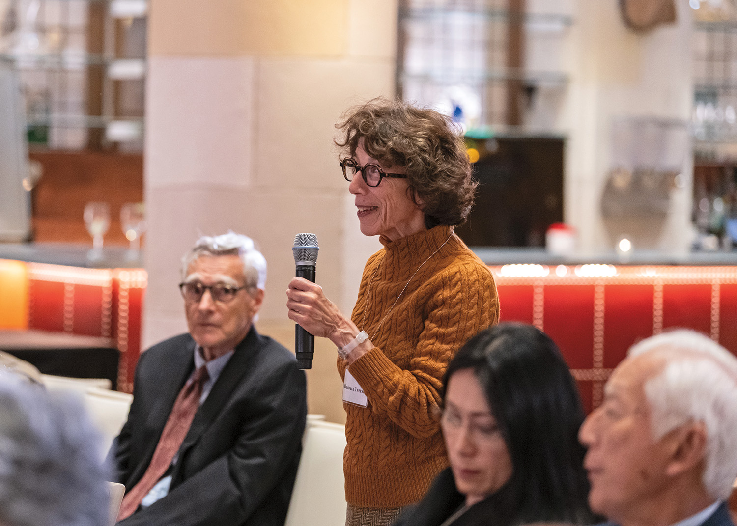 Barbara Tversky stands among seated attendees at event for the final report from the Commission on Reimaging Our Economy. Tversky holds a microphone and smiles. She has curly brown hair and pale skin. She wears glasses and a pumpkin-colored sweater.