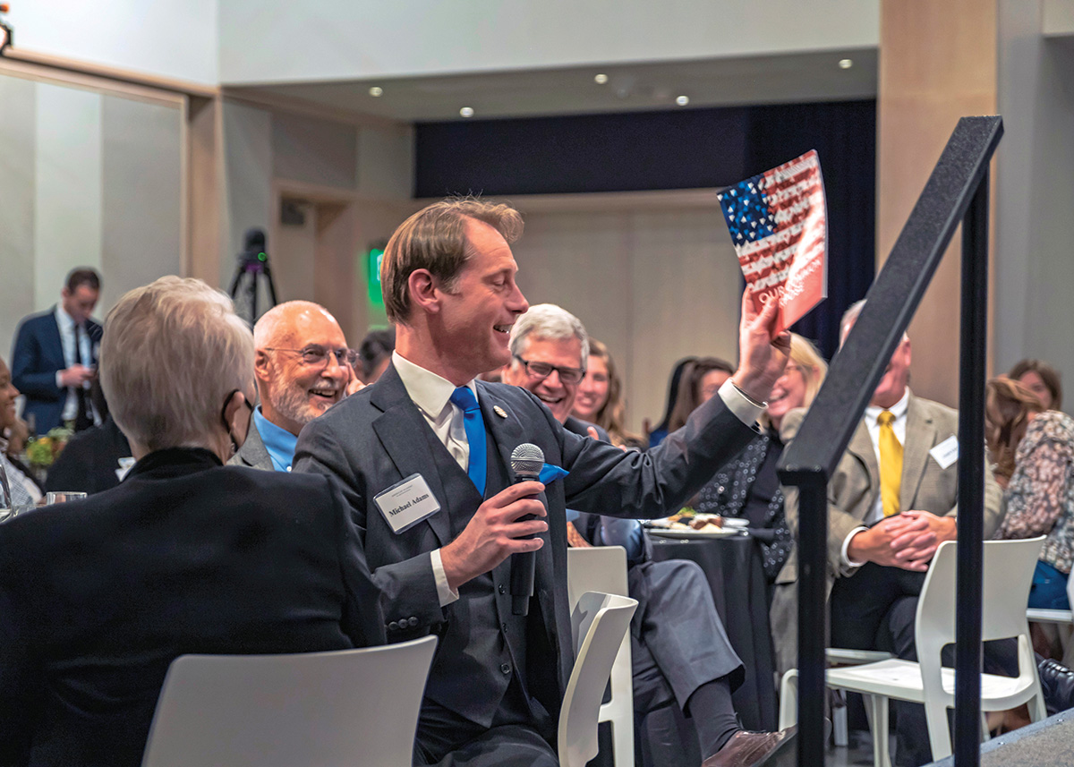Michael Adams sits among the attendees at an event to promote the Our Common Purpose report. Adams holds a microphone and a copy of Our Common Purpose. Adams has pale skin and short brown hair. He wears a suit and smiles at the other attendees.