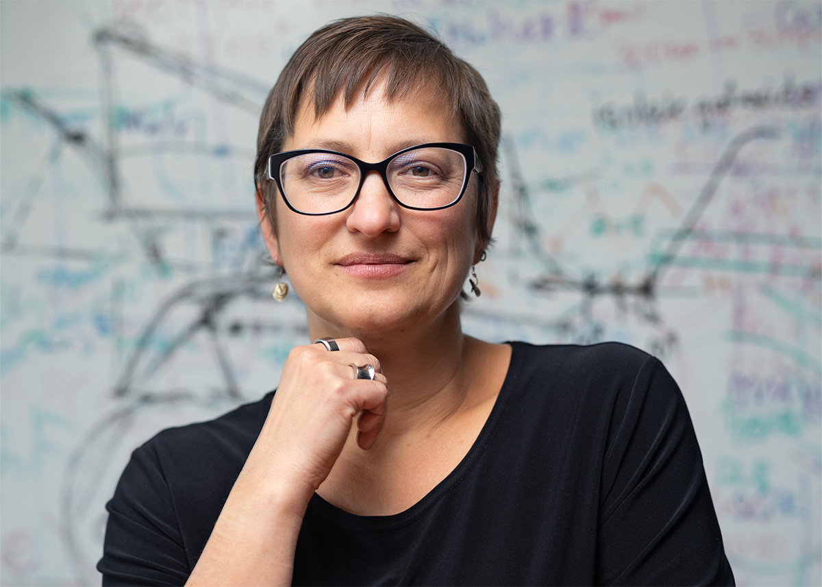 A photograph of a light-skinned individual with short brown hair. They are wearing black framed glasses and a black top. Their right hand rests under their chin. They are looking toward the viewer.