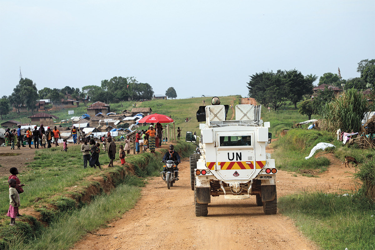 A motorcyclist rides their bike down a dirt road toward the viewer. A crowd of people gather in the field beside the road, and huts in various stages of construction stand on a hill that rises in the background.