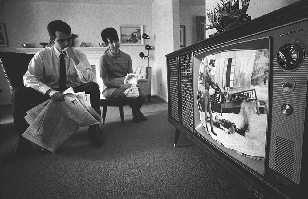 Black and white photo of a man and woman sitting in front of a television. The man is holding a newspaper.