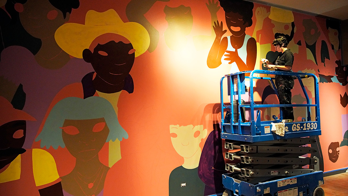 A person dressed in all black stands on a blue mechanical lift in front of a large mural featuring people with different hair, eye, and clothing colors and styles.