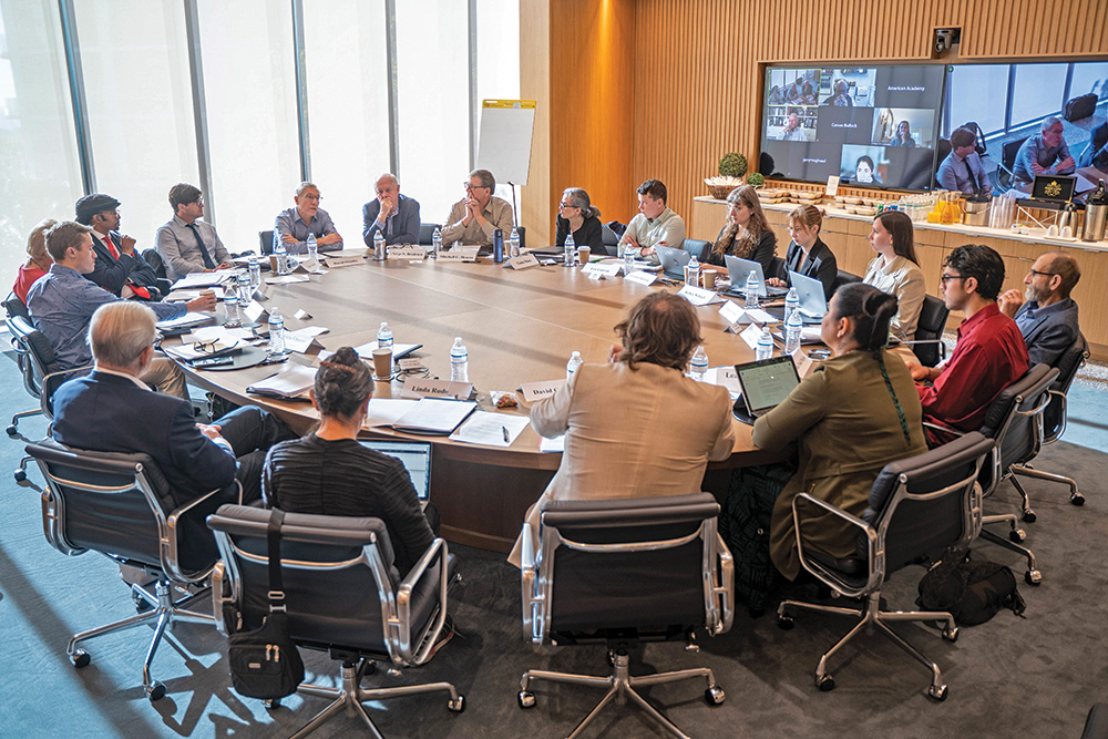 Eighteen people in business attire engage in discussion around a circular conference table. Eight people attend the meeting virtually, visible on a monitor behind the table.