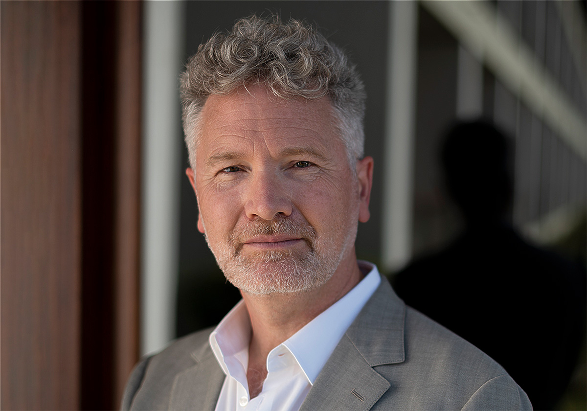 A light-skinned individual with curly gray hair looks toward the viewer. They are wearing a white business shirt and light gray blazer.