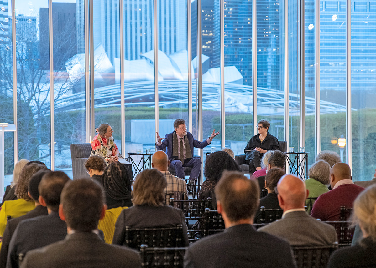 Three people are seated on a stage facing an audience. Oskar Eustis, the person in the middle, gestures broadly while talking. Laurie Patton, on his left, and Leah Dickerman, on his right, are listening.  