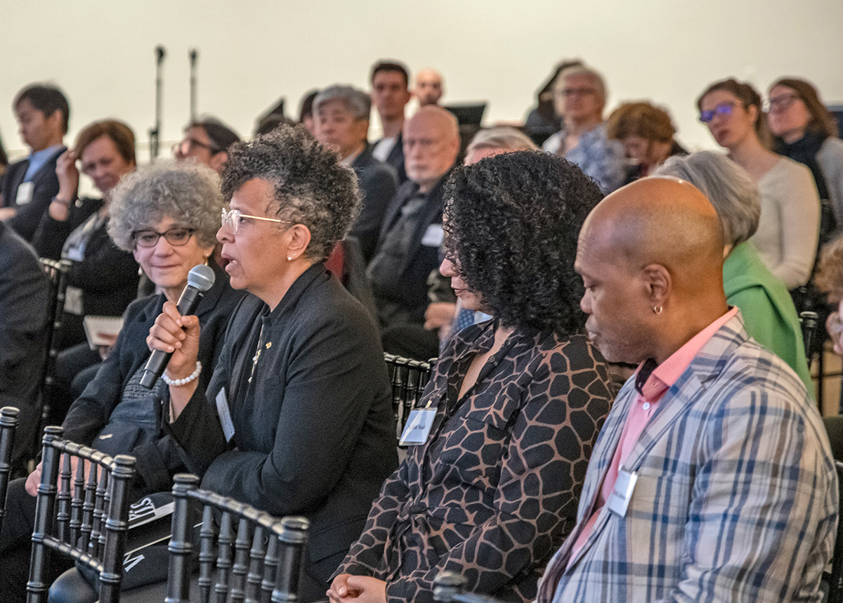 Sandra Jackson Dumont, a person with dark skin and dark curly hair, asks a question in the midst of a large audience looking in her direction. 